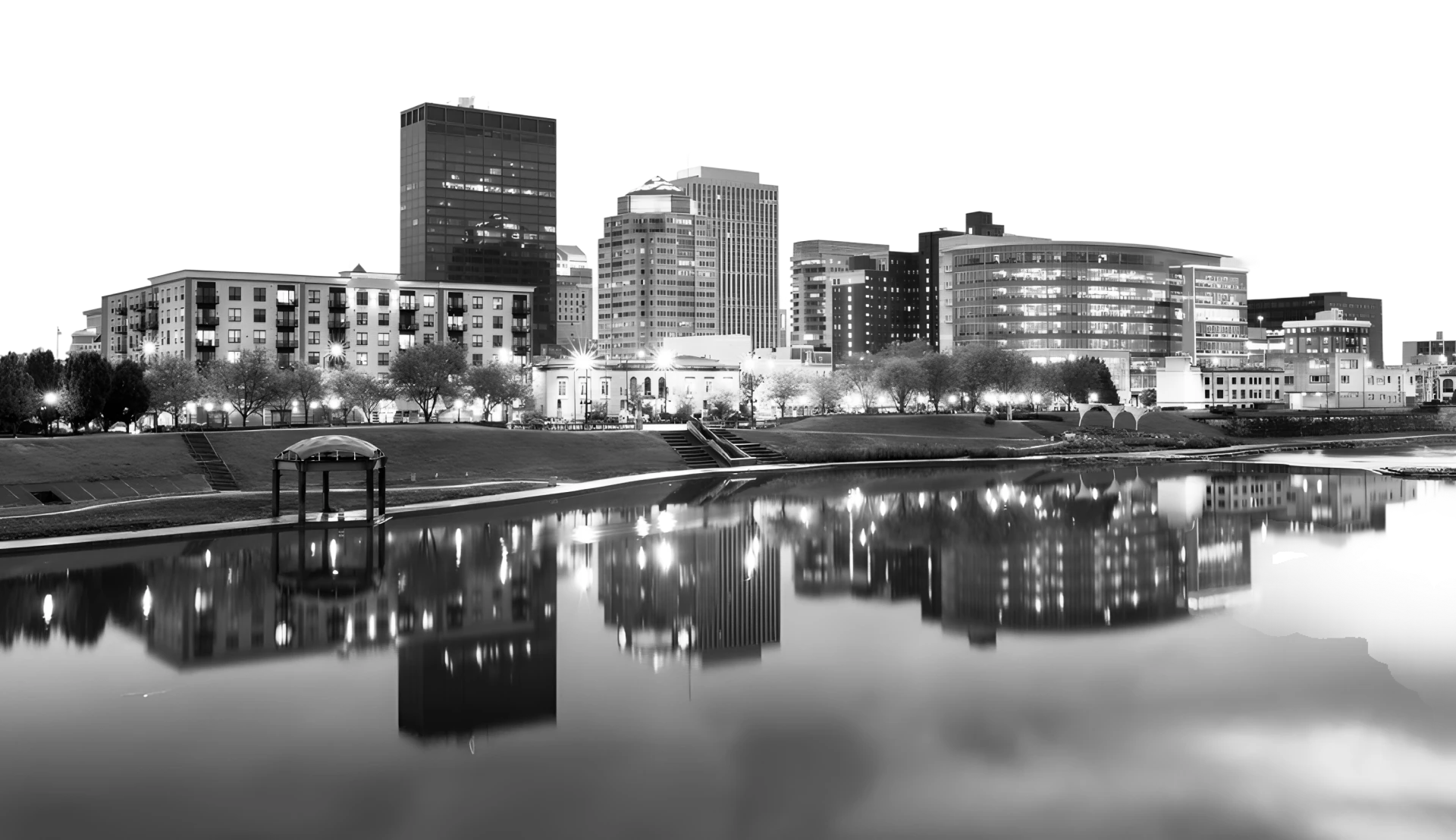 Black and white cityscape at night with illuminated buildings reflected in a calm river, a park, and a gazebo.