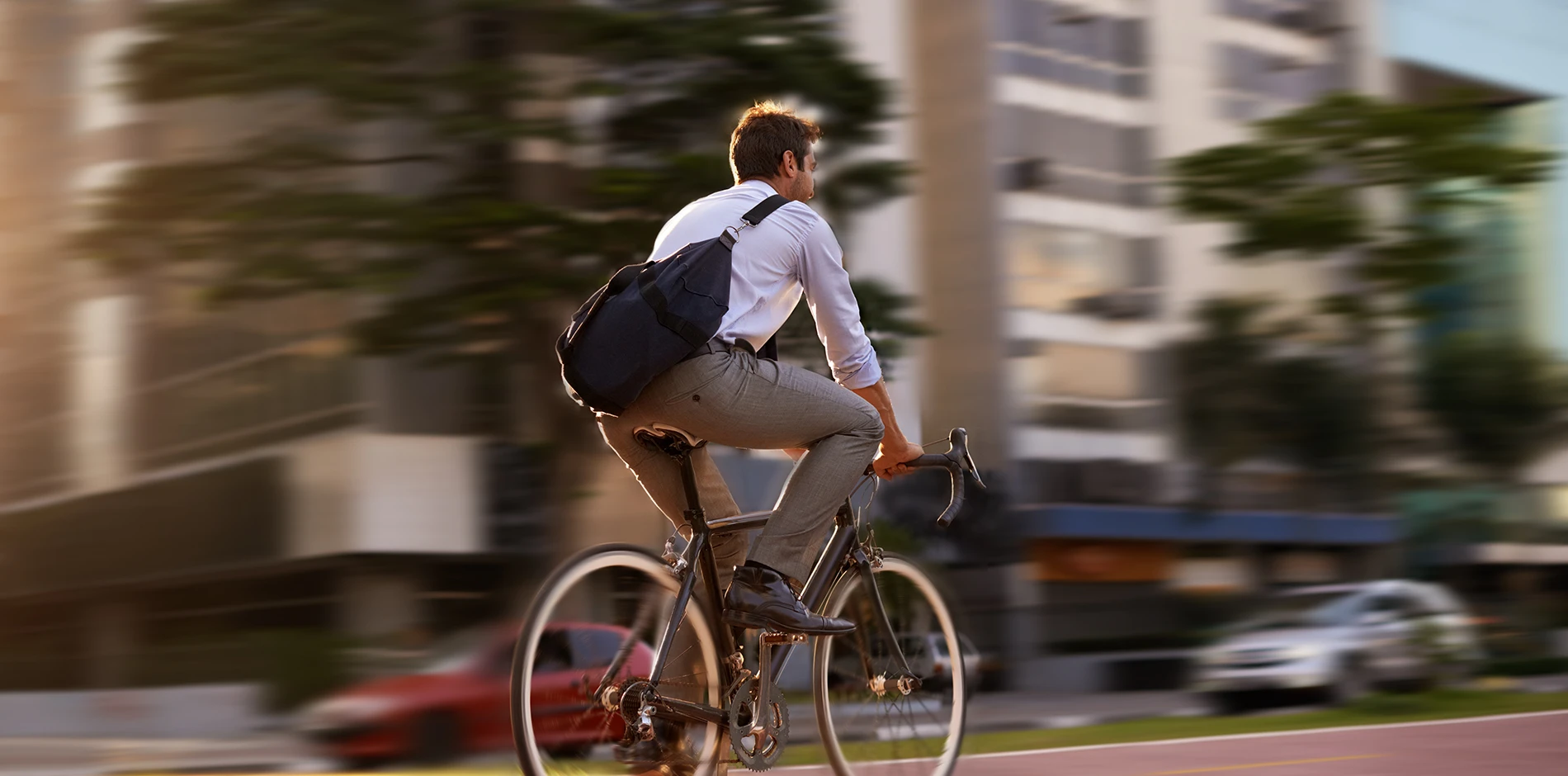 Man in white shirt, gray trousers, and shoulder bag cycles a black bike on a blurred city street.