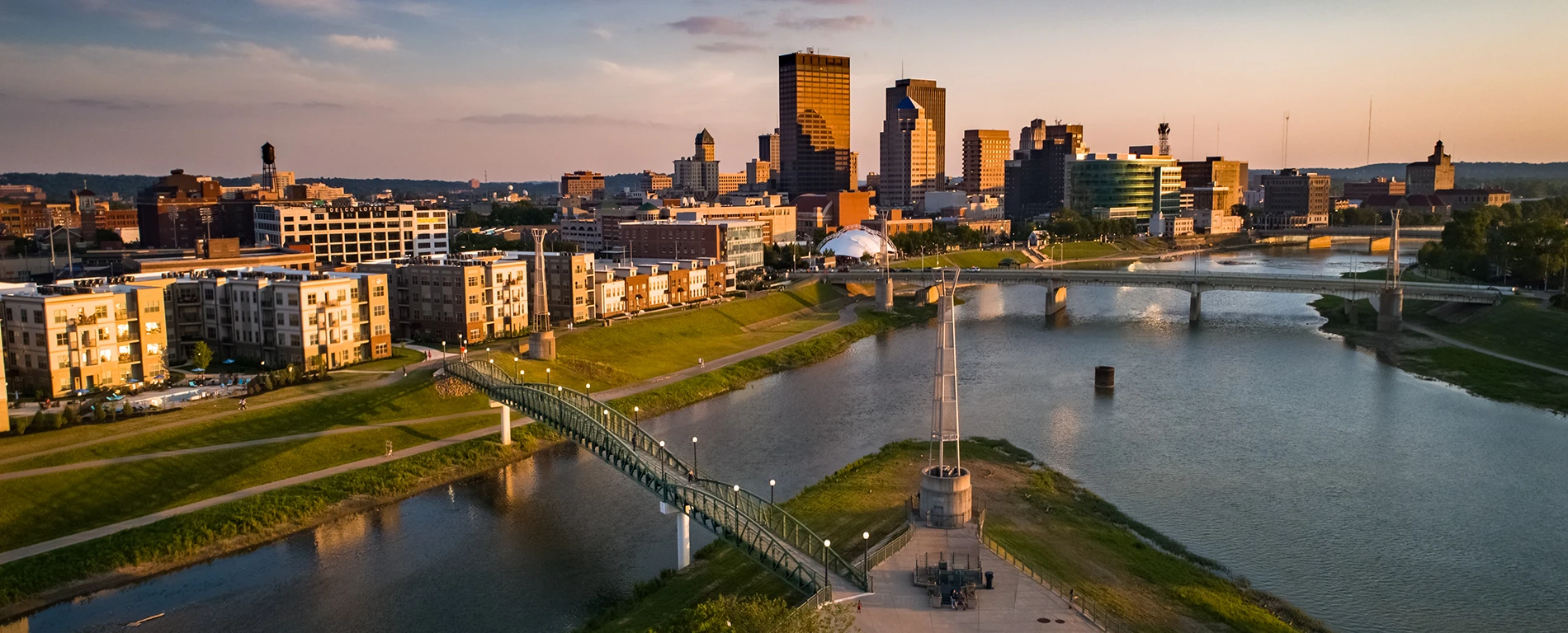 Aerial view of a vibrant cityscape with a river, multiple bridges, modern buildings, and green riverbanks at sunset.