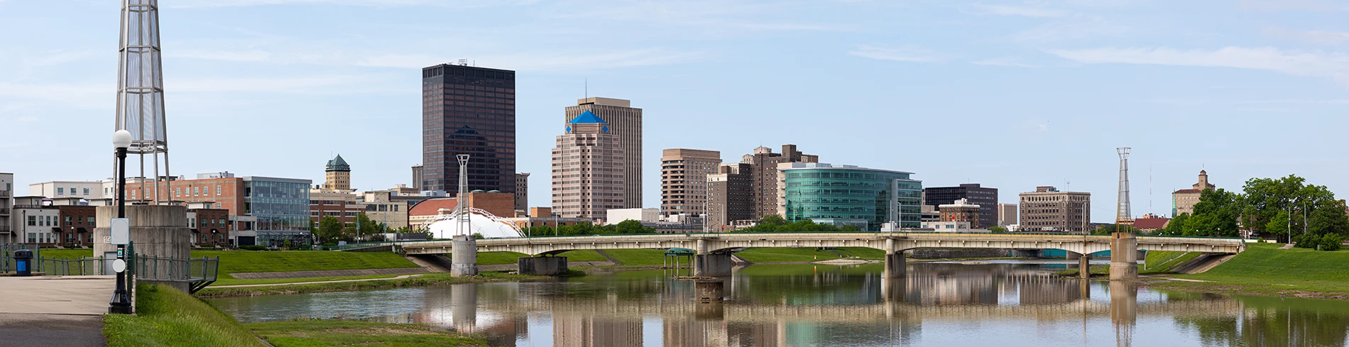 Panoramic city skyline with diverse buildings reflected in a calm river, spanned by a bridge, and green banks.