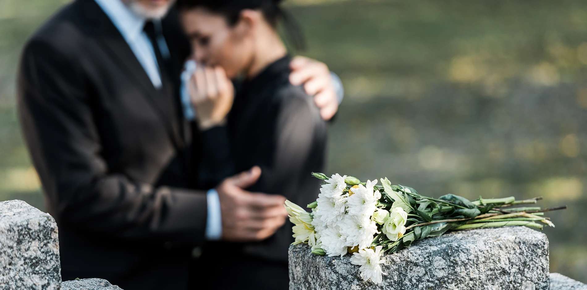 White flowers on a gravestone, a man comforts a crying woman in black mourning clothes.