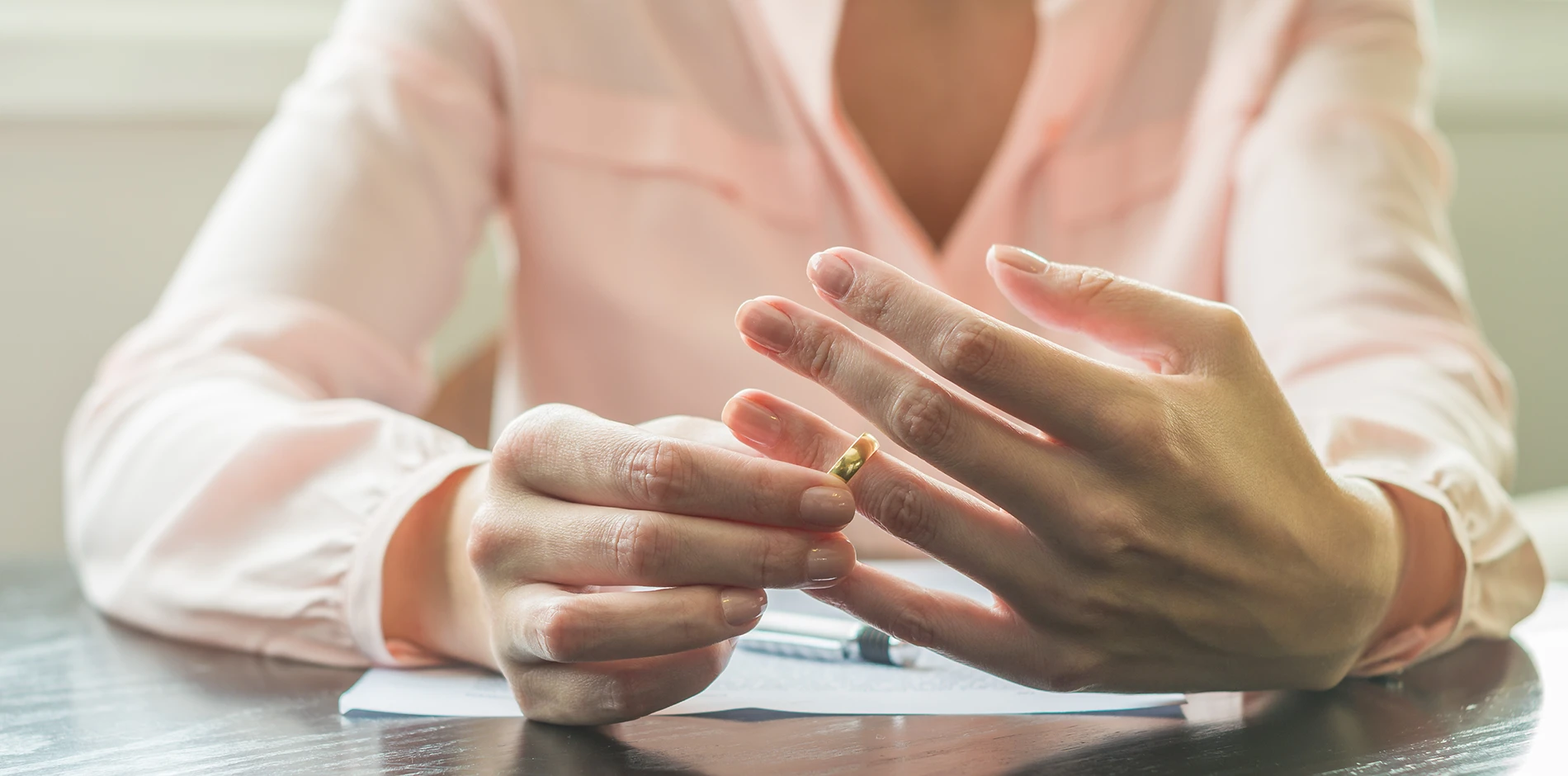 A woman in a pink shirt removes a golden wedding ring from her finger above papers on a desk.