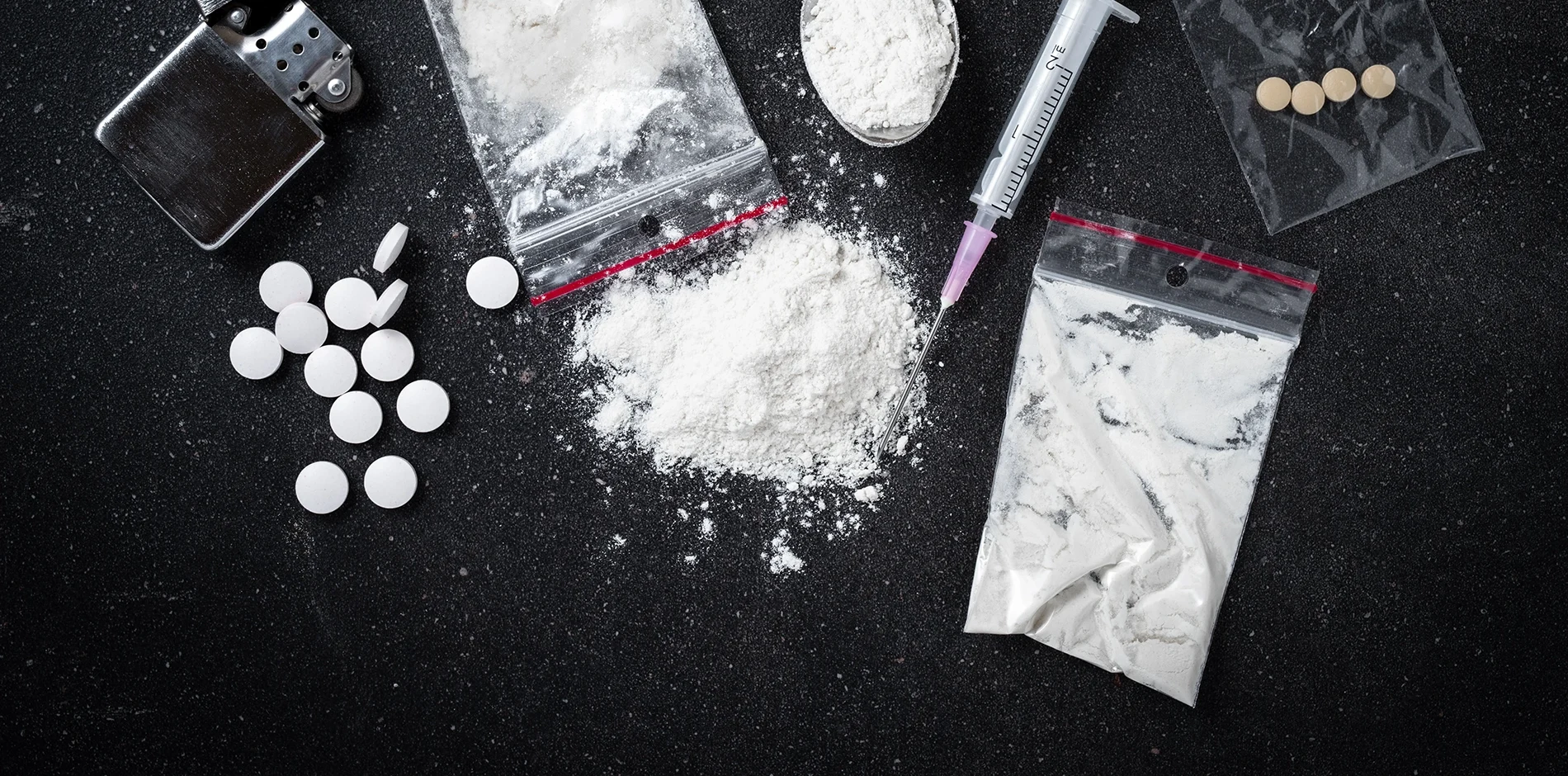 White powder, multiple pills, a syringe, and a lighter scattered on a dark surface.
