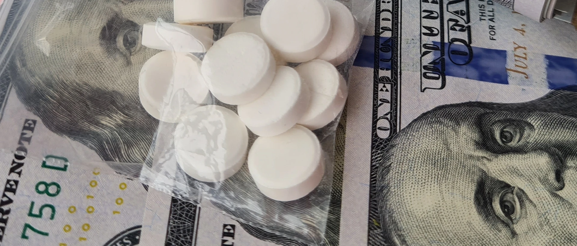 White, round tablets in a clear plastic bag rest on US $100 bills.