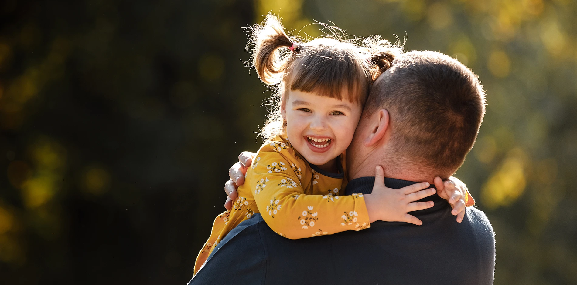 Smiling girl with pigtails embracing her father from behind, backlit by sunlight in nature.