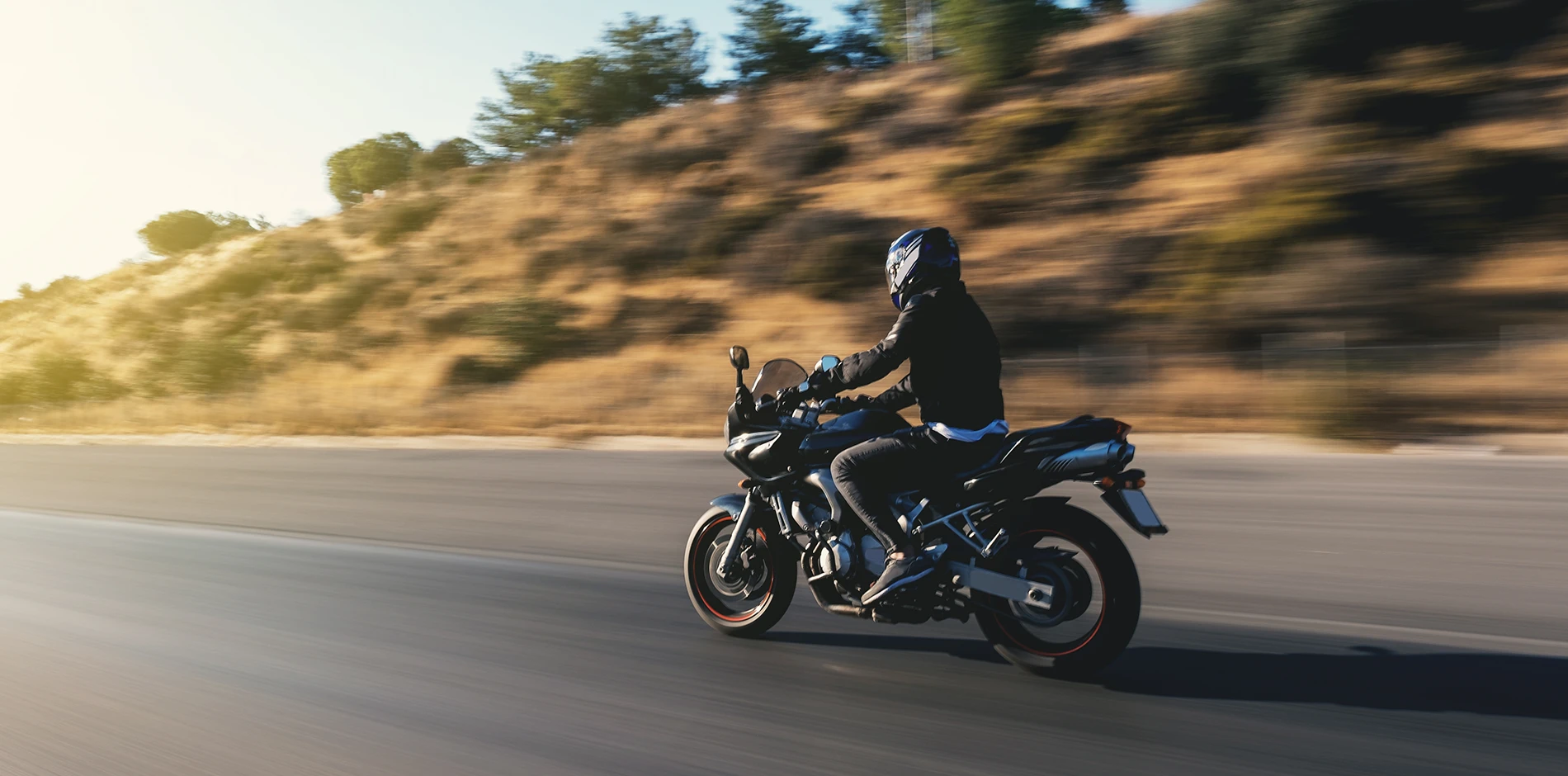 Rider in a blue helmet speeds on a dark motorcycle along a road beside a blurred, sunny hill.