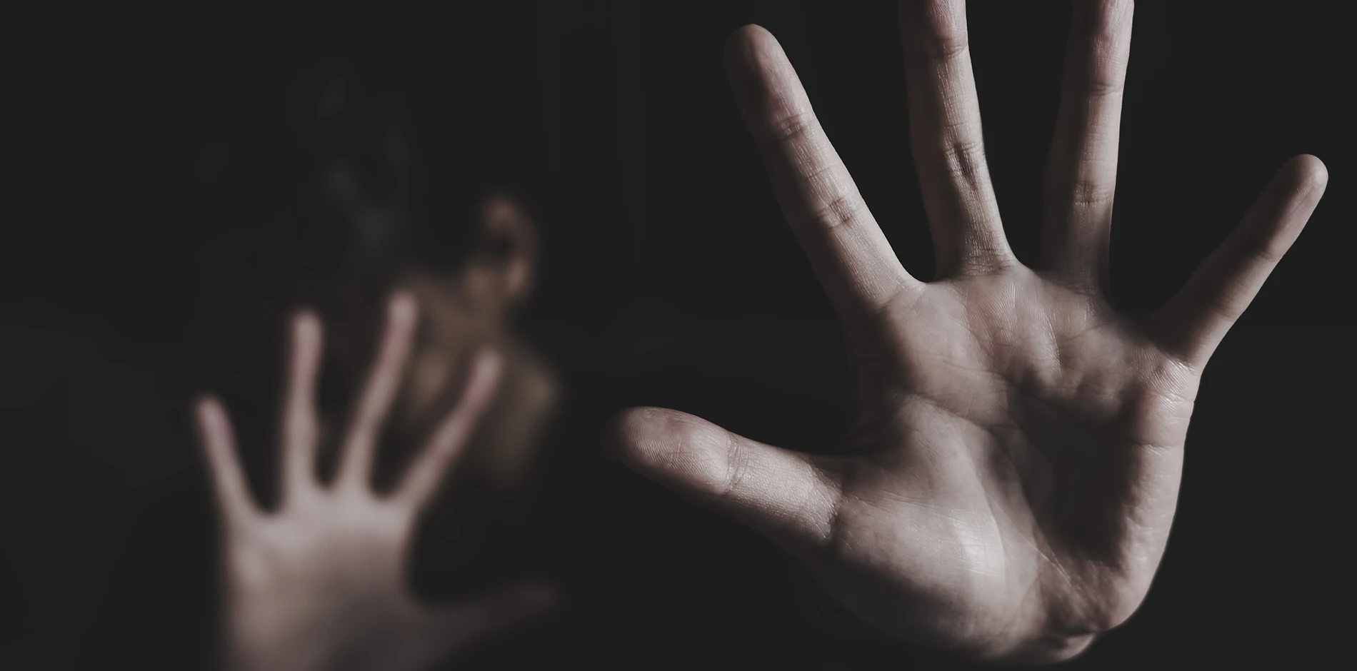 Clear hand with palm out, fingers spread, extended towards viewer; blurry person and hand in dark background.