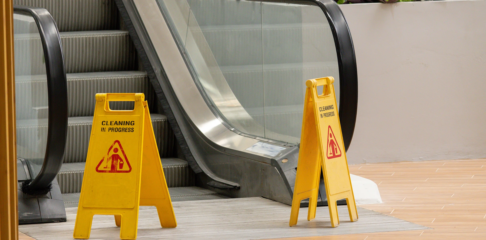Two yellow Cleaning in Progress slip hazard signs with a mopping pictogram stand by an escalator.
