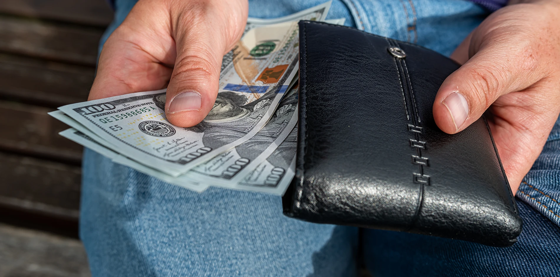 Hands hold a black wallet with several hundred-dollar bills, against blue jeans.