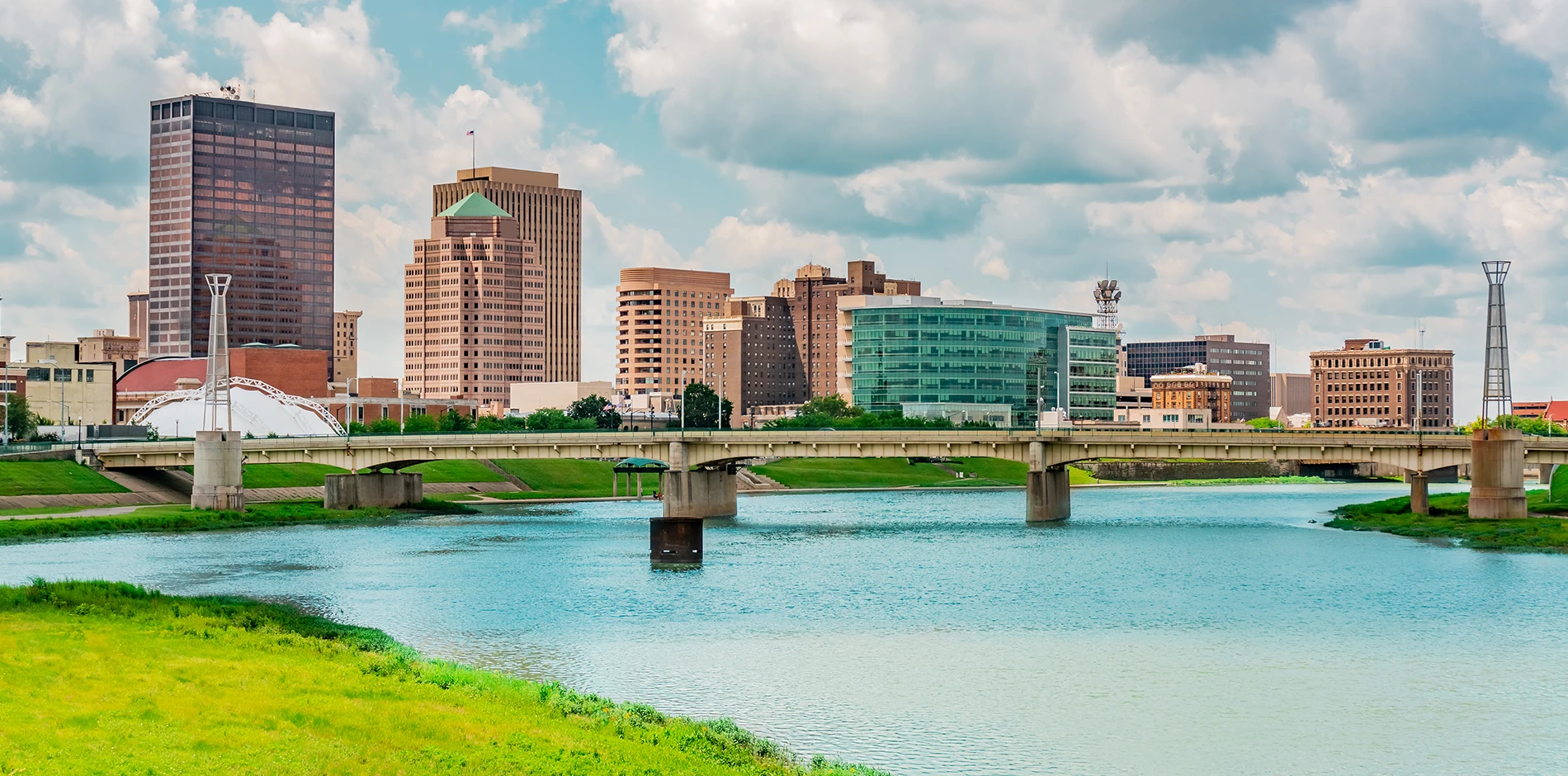 Skyline of diverse tall buildings with a bridge spanning a wide river and green banks under a cloudy sky.