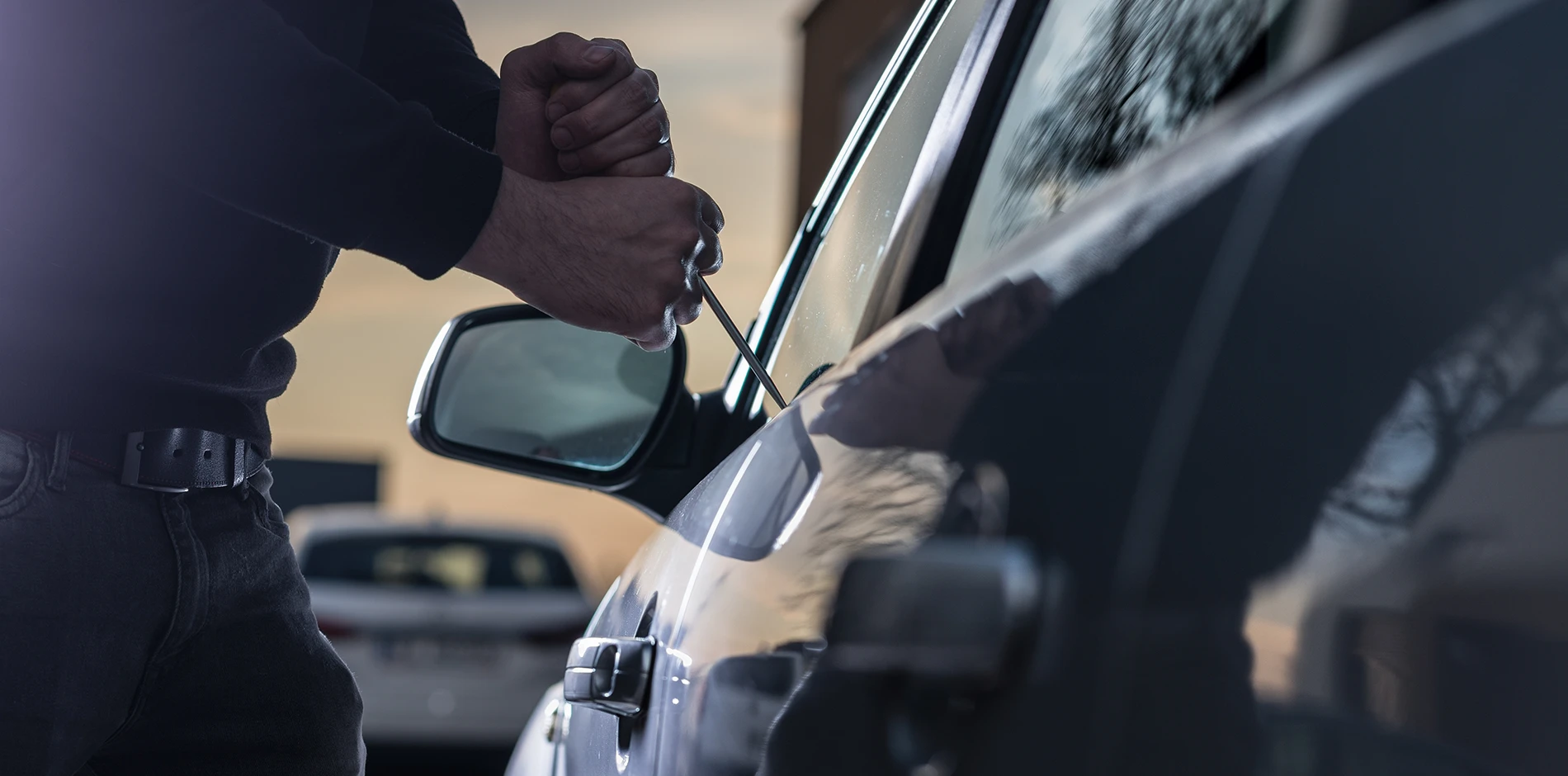 Theft uses a screwdriver to pry open the top of a dark car's driver side window.