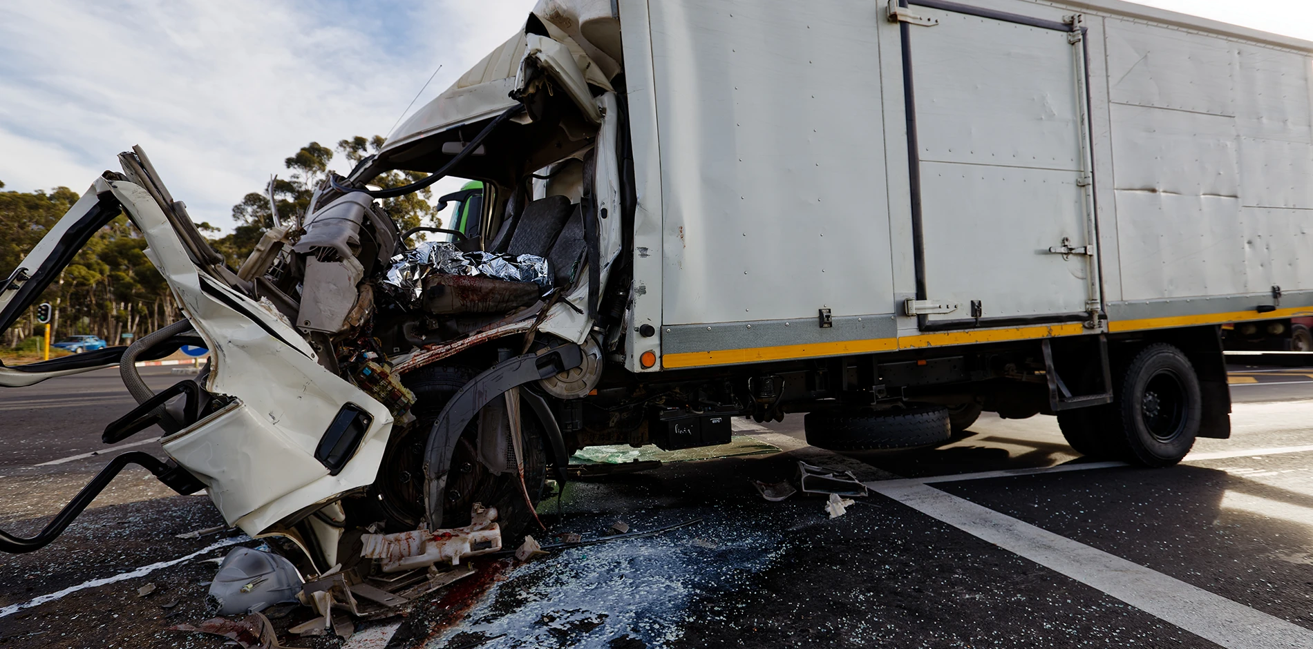 Mangled white vehicle and damaged white box truck on an asphalt road, surrounded by debris and blood.