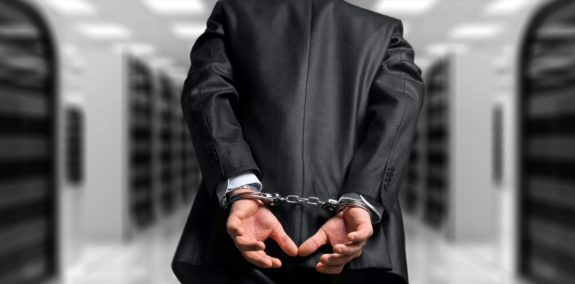 A white-collar individual in a dark suit with hands cuffed behind their back in a blurry office setting.